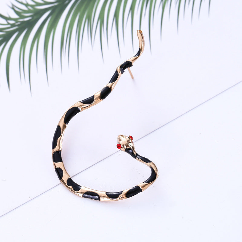 A gold-colored snake-shaped earring with a dripping oil design, displayed against a white background with a green leafy element to the side.